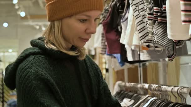 Bir kız bir yeşil kazak ve sarı şapka şeyler mağazası aracılığıyla yürüyor ve ne satın almak için seçer. Askıları şeylerde dokunur ve fiyat etiketleri görünüyor — Stok video