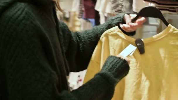 Una chica con un suéter verde y un sombrero amarillo camina a través de una tienda de cosas y elige qué comprar. Toca cosas en perchas y mira etiquetas de precios — Vídeos de Stock