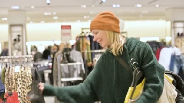 Une fille dans un pull vert et un chapeau jaune marche à travers un magasin de choses et choisit ce qu'il faut acheter. Touche les choses sur les cintres et regarde les étiquettes de prix — Video