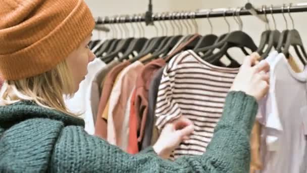 Дівчина в зеленому светрі і жовтий капелюх проходить через магазин речей і вибирає, що купити. Торкається речей на вішалках і дивиться на цінові теги — стокове відео