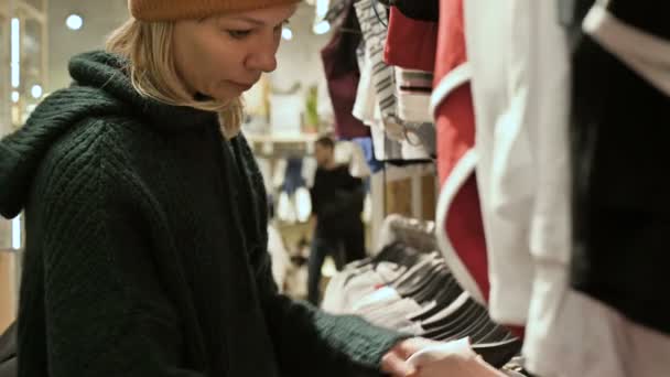 Una chica con un suéter verde y un sombrero amarillo camina a través de una tienda de cosas y elige qué comprar. Toca cosas en perchas y mira etiquetas de precios — Vídeos de Stock