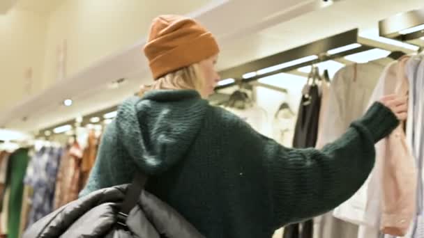 Een meisje in een groene trui en een gele hoed begeleidt bij een winkel van dingen en kiest wat te kopen. Raakt dingen op hangers en kijkt naar de prijskaartjes — Stockvideo