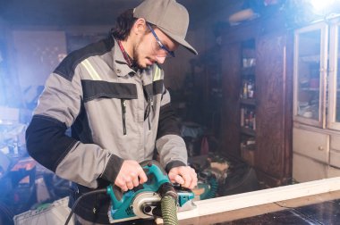 Genç sakallı bir elektrikli uçak talaş emme ile çalışan bir marangoz. Tesviye ve ahşap çubukları zımpara
