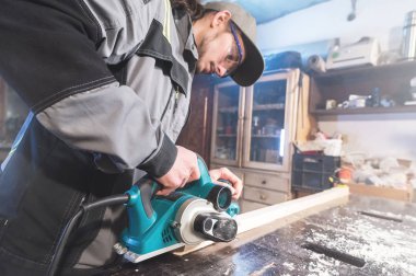 Genç sakallı bir elektrikli uçak talaş emme olmadan çalışan bir marangoz. Tesviye ve ahşap çubukları zımpara. Düşük geniş açı