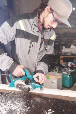 Sakallı genç marangoz elektrikli bir uçakla talaş emme olmadan çalışır. Tesviye ve ahşap kirişler zımpara