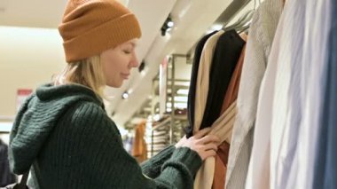 Bir kız bir yeşil kazak ve sarı şapka şeyler mağazası aracılığıyla yürüyor ve ne satın almak için seçer. Askıları şeylerde dokunur ve fiyat etiketleri görünüyor