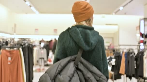 Vista dal retro ragazza shopping in un maglione e cappello cammina attraverso un negozio di abbigliamento. Telecamera live — Video Stock