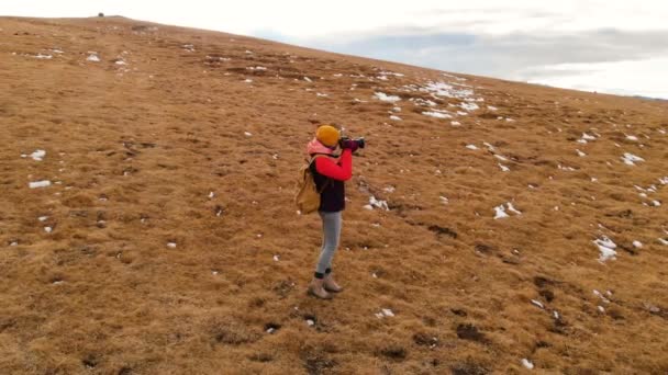 Hava görünümü. Kız gezgin bir sırt çantası ve gün batımında Dağları arka planı sarı çim ile Yamaç ve bulutlu bir plato boyunca yürüyüş güneş gözlüğü ile şapkalı bir kamera ile — Stok video