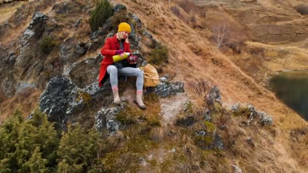 Vista aérea de una niña sentada en una roca de un promontorio en la orilla del lago vierte té de un termo — Vídeos de Stock