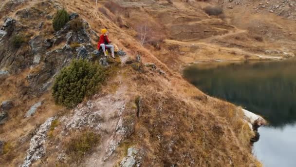 Vista aérea de una chica sentada en una roca de un cabo en la orilla ozerapet té disfrutando de la vista en las montañas — Vídeos de Stock