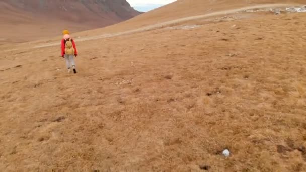 Luft nach oben. Reisendes Mädchen mit Kamera, Hut, Rucksack und Sonnenbrille, das den Hang entlang geht, mit gelbem Gras vor dem Hintergrund von Bergen und einem Plateau mit Wolken bei Sonnenuntergang — Stockvideo