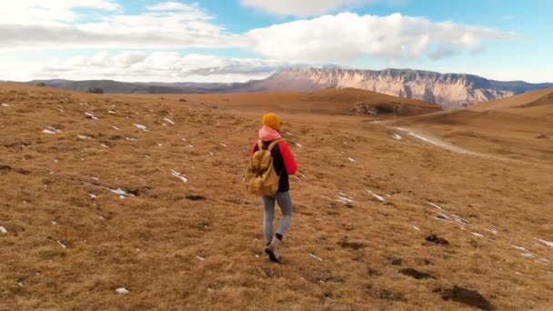 Vista aérea. Chica viajera con una cámara en un sombrero con una mochila y gafas de sol caminando por la ladera con hierba amarilla sobre el fondo de las montañas y una meseta con nubes al atardecer — Vídeo de stock