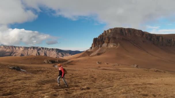 Vista aérea. Viajante menina com uma câmera no pescoço andando ao longo da encosta com grama amarela contra o pano de fundo das montanhas e um planalto com nuvens ao pôr do sol — Vídeo de Stock