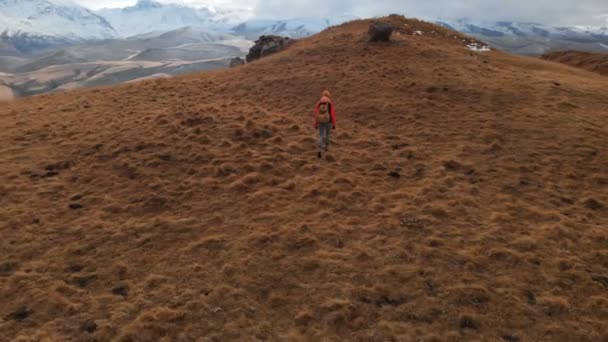 Vista aérea. Chica viajera con una cámara en el cuello caminando por la ladera con hierba amarilla contra el telón de fondo de las montañas y una meseta con nubes al atardecer — Vídeos de Stock