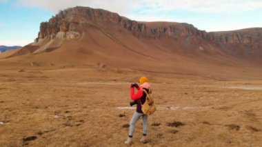 Hava görünümü. Bir gezgin kız onun etrafında bir kamera ile boyun ve yamaç arka plan dağlar ve bulutlu bir plato günbatımında yürüyor. Kız resimleri telefonda alır.