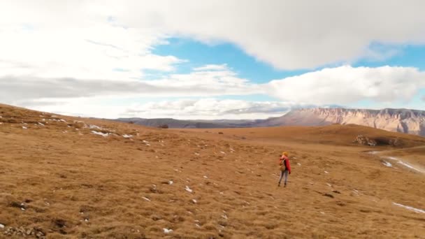 어 볼 수 있습니다. 여자 여행자 배낭과 일몰에 산맥의 배경에 대 한 노란 잔디 슬로프와 구름과 대 지를 따라 산책 하는 선글라스와 모자에 카메라와 함께 — 비디오