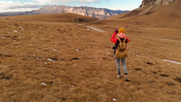 Luftvy. En resenär flicka med en kamera runt henne hals och promenerar längs sluttningen en bakgrund av berg och en platå med moln i solnedgången. Flickan tar bilder på telefonen — Stockvideo