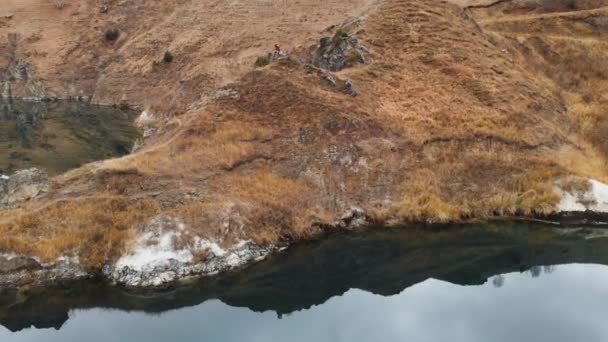 Αεροφωτογραφία του ένα κορίτσι που κάθεται σε έναν βράχο σε μια λίμνη που παίρνει μια φωτογραφία του τοπίου στο τηλέφωνο και να πυροβολήσει ένα βίντεο. Ταξίδια βίντεο — Αρχείο Βίντεο