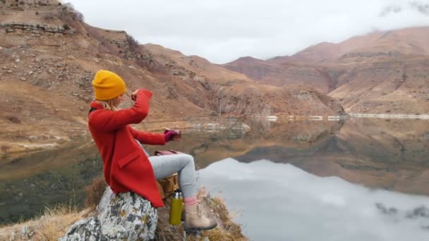 Egy lány ül egy szikla egy tóparti, hogy fog egy fénykép, a táj, a telefon, és lő egy videó a légifelvételek. Utazási videó — Stock videók