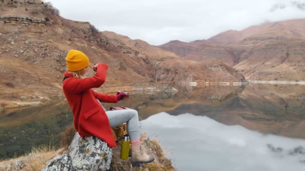 Vista aérea de uma menina sentada em uma rocha em um lago que tira uma foto da paisagem no telefone e filma um vídeo. Vídeo de viagem — Vídeo de Stock