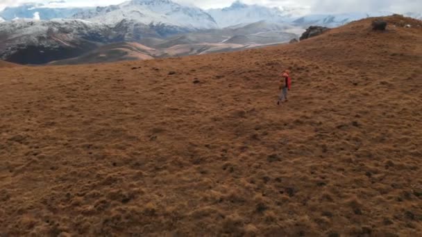 Vista aérea. Chica viajera con una cámara en el cuello caminando por la ladera con hierba amarilla contra el telón de fondo de las montañas y una meseta con nubes al atardecer — Vídeo de stock