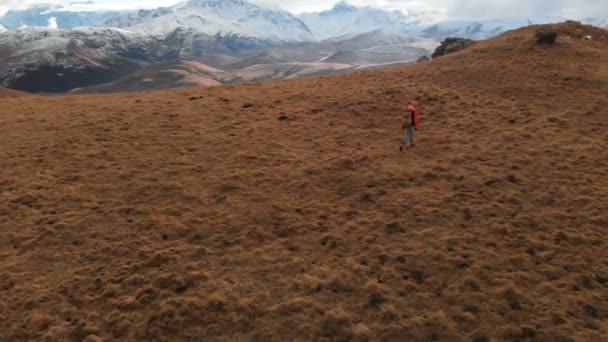 Vista aérea. Chica viajera con una cámara en el cuello caminando por la ladera con hierba amarilla contra el telón de fondo de las montañas y una meseta con nubes al atardecer — Vídeos de Stock