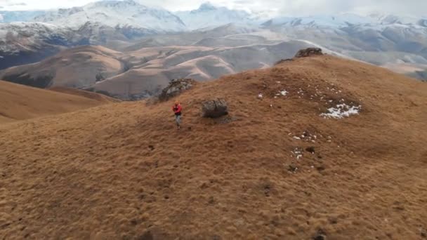 Vista aérea. Chica viajera con una cámara en el cuello caminando por la ladera con hierba amarilla contra el telón de fondo de las montañas y una meseta con nubes al atardecer — Vídeos de Stock
