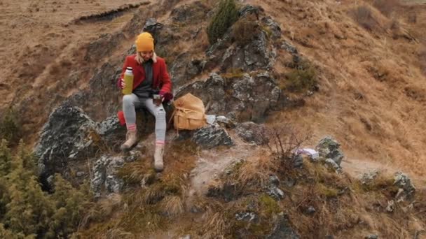 Vista aérea de una niña sentada en una roca de un promontorio en la orilla del lago vierte té de un termo — Vídeos de Stock