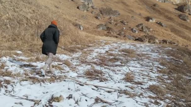 Chica turista en una chaqueta y una gorra camina a lo largo del camino a principios de invierno. hierba amarilla y trozos de nieve en un día soleado. Vista aérea — Vídeo de stock