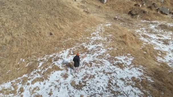 Kız turist aşağı bir ceket ve bir kap içinde yol boyunca erken kış aylarında yürür. Sarı çim ve güneşli bir günde kar parçaları. Havadan görünümü — Stok video