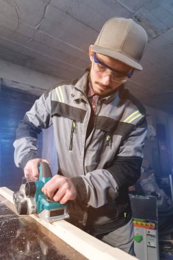 Genç sakallı bir elektrikli uçak talaş emme olmadan çalışan bir marangoz. Tesviye ve ahşap çubukları zımpara. Düşük geniş açı