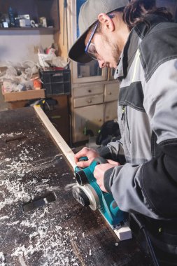 Genç sakallı bir elektrikli uçak talaş emme olmadan çalışan bir marangoz. Tesviye ve ahşap çubukları zımpara. Yüksek geniş açı