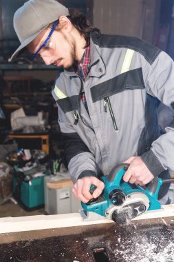 Sakallı genç marangoz elektrikli bir uçakla talaş emme olmadan çalışır. Tesviye ve ahşap kirişler zımpara