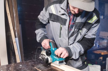 Sakallı genç marangoz elektrikli bir uçakla talaş emme olmadan çalışır. Tesviye ve ahşap kirişler zımpara