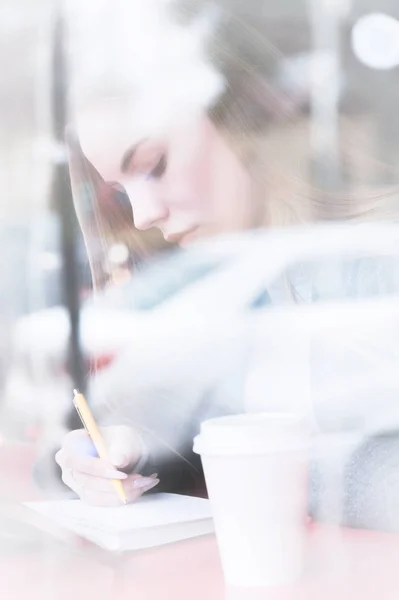 Portrét dívky atraktivní mladý novinář s pero a Poznámkový blok v kavárně za vitríny. Non kontrastní zobrazení prostřednictvím okna skla reflexe — Stock fotografie