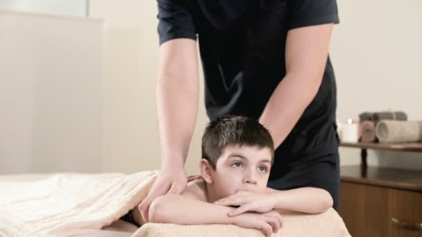 Мужчина массажист-физиотерапевт делает лечебный расслабляющий массаж маленькому мальчику, лежащему на массажной кровати. Массаж рук и плеч — стоковое видео