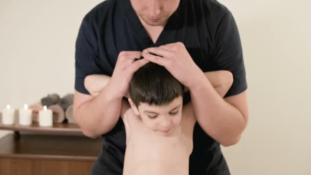 Jovem massagista fisioterapeuta do sexo masculino fazendo alongamento terapêutico das costas e coluna vertebral para uma criança pequena em uma sala de massagem — Vídeo de Stock