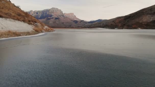 Luchtfoto van een lage vlucht over een bevroren meer in de winter, omringd door de bergen van de Kaukasus na zonsondergang op het blauwe uur — Stockvideo