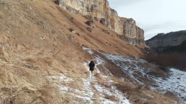 ダウン ジャケットと帽子の女の子観光は、岩の多い峡谷に初冬のパスに沿って歩きます。黄色の草や晴れた日の雪の部分。航空写真ビュー — ストック動画