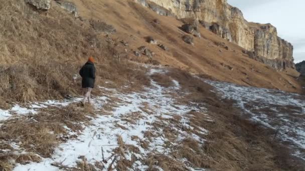 Chica turista en una chaqueta y sombrero camina a lo largo del camino a principios de invierno en un barranco rocoso. hierba amarilla y trozos de nieve en un día soleado. Vista aérea — Vídeo de stock