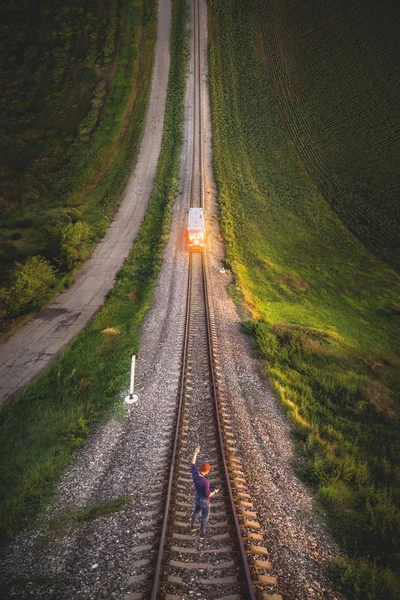 Aerial view A male hipster in a cap holding up a hand with a gesture Victory is standing on rails in front of an approaching train. Reverse vertical panorama in the style of Inception — Stock Photo, Image