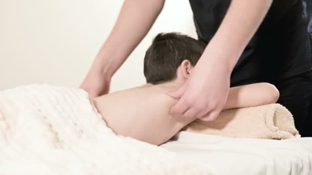 Мужчина массажист-физиотерапевт делает лечебный расслабляющий массаж маленькому улыбающемуся мальчику, лежащему на массажной кровати. Массаж поясницы и спины — стоковое видео