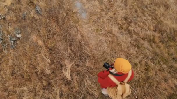 Крупный план Вид сверху с высоты птичьего полета на девушку-фотографа в жёлтой шляпе с рюкзаком, идущим вдоль горной тропы. Видео путешествия — стоковое видео