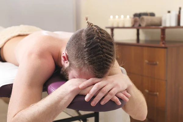 Male hipster with braided trendy hair and shaved temples lying on a massage bunk down face resting and waiting for physical therapy and massage — Stock Photo, Image