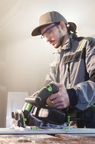 Портрет молодого столяра с ручной циркулярной пилой в руках рабочего в домашней мастерской. Открываю свой бизнес. Мбаппе
