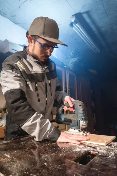 Gri tulum ve gözlük mesleği sakallı genç adam bir marangoz bir elektrikli testere onun ev atölye olarak çalışıyor. Kesme ahşap parçaları