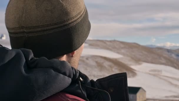 在冬天的山上, 一个穿着温暖夹克、戴着帽子的白人男子的特写镜头后视了看日落时的山脉。头部旋转 — 图库视频影像