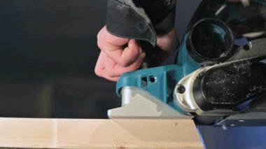 Büyük yakın çekim. Bir marangoz, elektrikli düzlemde ahşap bir tahta kesiyor. Toz parçacıklarının ve testere bıçağının yavaş hareketi