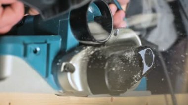 Büyük yakın çekim. Bir marangoz, elektrikli düzlemde ahşap bir tahta kesiyor. Toz parçacıklarının ve testere bıçağının yavaş hareketi