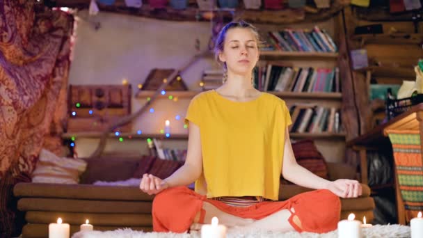 Una hermosa joven con los ojos cerrados en ropa suelta se sienta en una sala de manualidades en una piel de piel blanca en una posición de loto rodeado de velas. Meditación crecimiento espiritual y cuidado de su propia salud — Vídeos de Stock